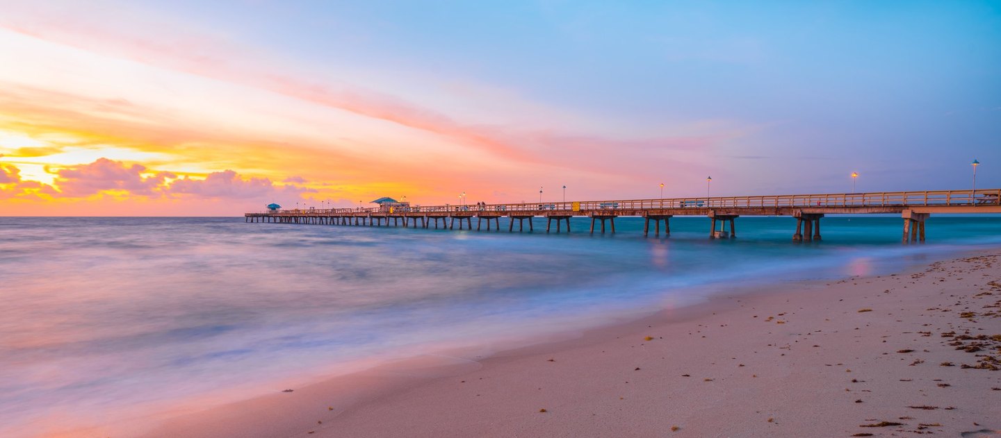 A long pier with glowing lamps extends over softly blurred blue waves beside a sandy beach beneath a pink, gold, and blue sky.