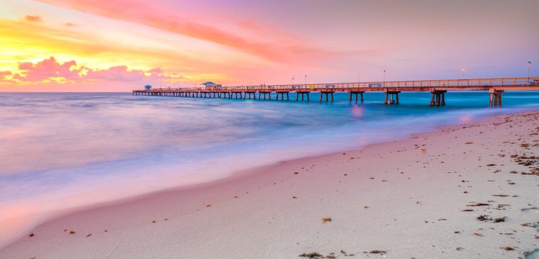 A long pier stretches across turquoise water beneath a pink and golden sky, with softly blurred surf washing onto a sandy beach scattered with seaweed.