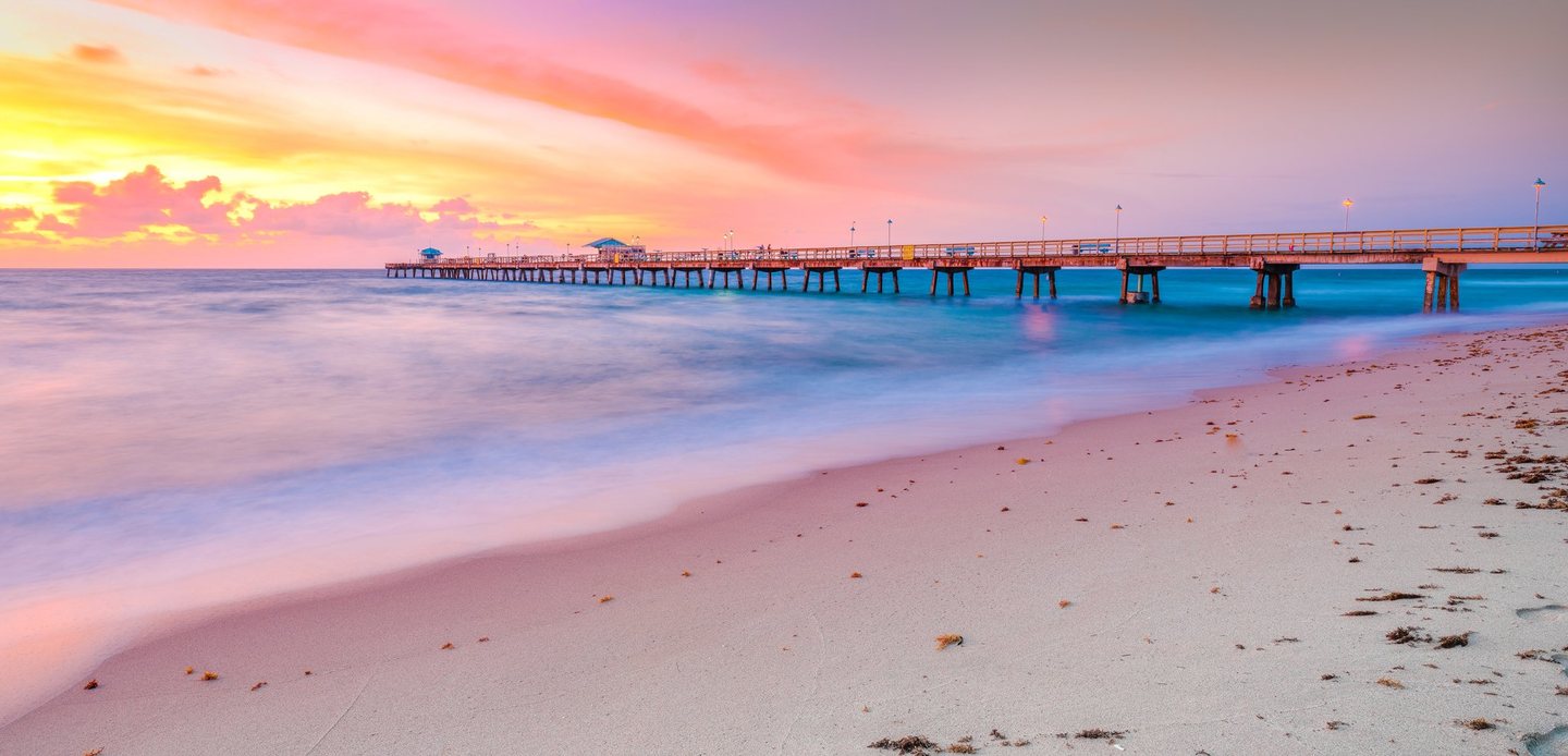 A long pier stretches across turquoise water beneath a pink and golden sky, with softly blurred surf washing onto a sandy beach scattered with seaweed.