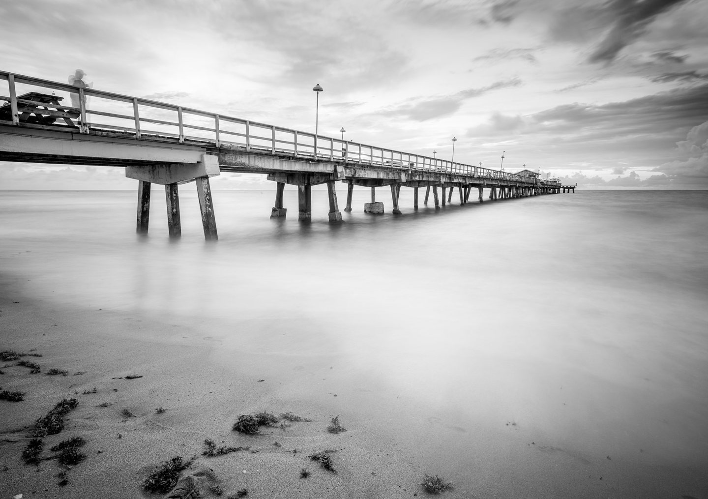 Black-and-white photograph of a lamp-lined pier extending over smooth ocean water beneath streaked clouds, with seaweed scattered across the sandy foreground.