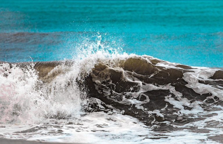 A dark ocean wave breaks into white spray and trailing foam against a broad expanse of turquoise water.