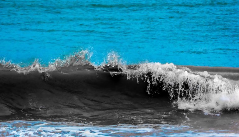 A dark ocean wave breaks into white spray against vivid turquoise water, with swirling foam along the foreground.