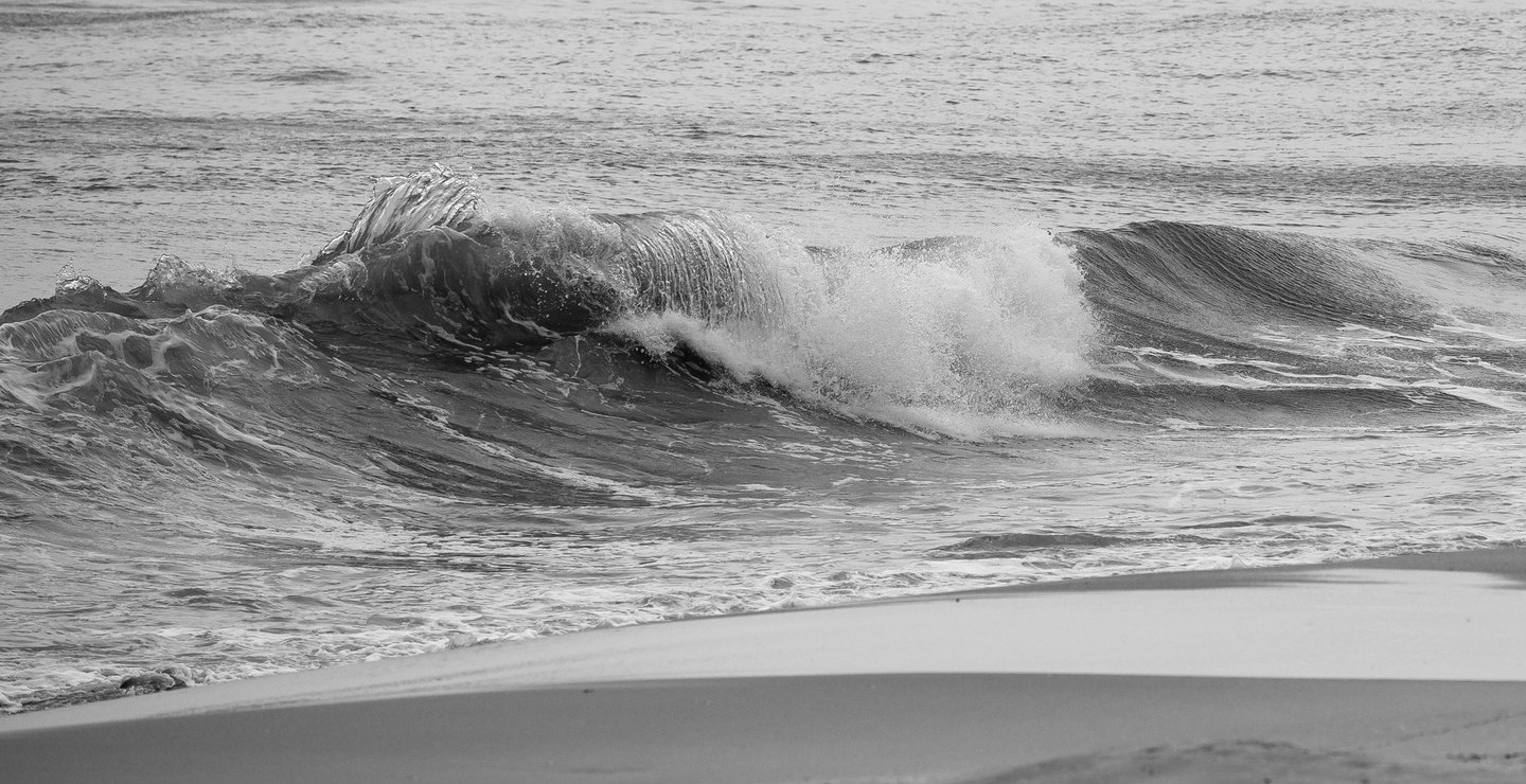 Black-and-white photograph of an ocean wave breaking into white spray beside a smooth, curving stretch of sandy beach.