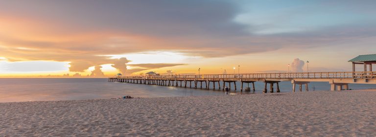 A long pier with illuminated lamps extends over calm water beside a sandy beach beneath orange, gold, and blue-gray clouds.