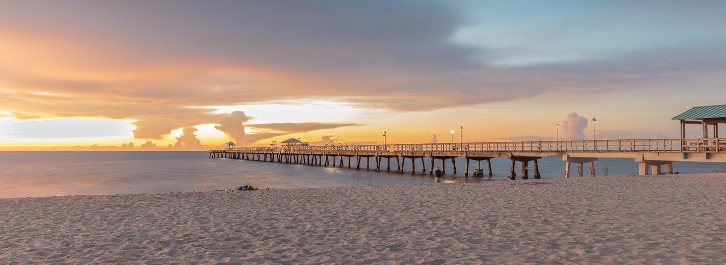 A long pier with illuminated lamps extends over calm water beside a sandy beach beneath orange, gold, and blue-gray clouds.
