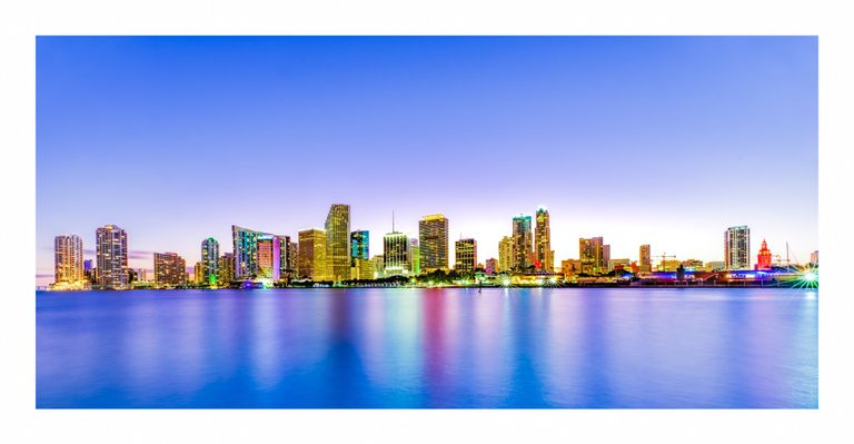 Miami’s illuminated waterfront skyline stretches beneath a clear blue and lavender twilight sky, with colorful reflections across the bay.