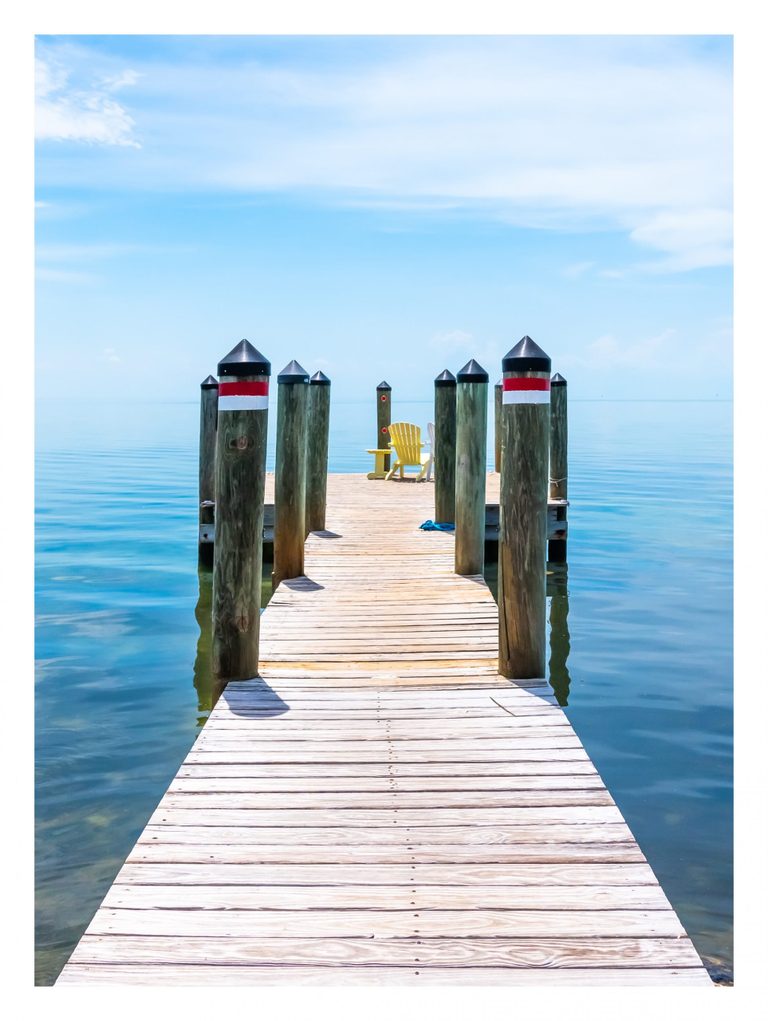 A sunlit wooden dock lined with capped timber posts extends across calm blue water toward a yellow chair and a partially visible white chair beneath a lightly clouded sky.
