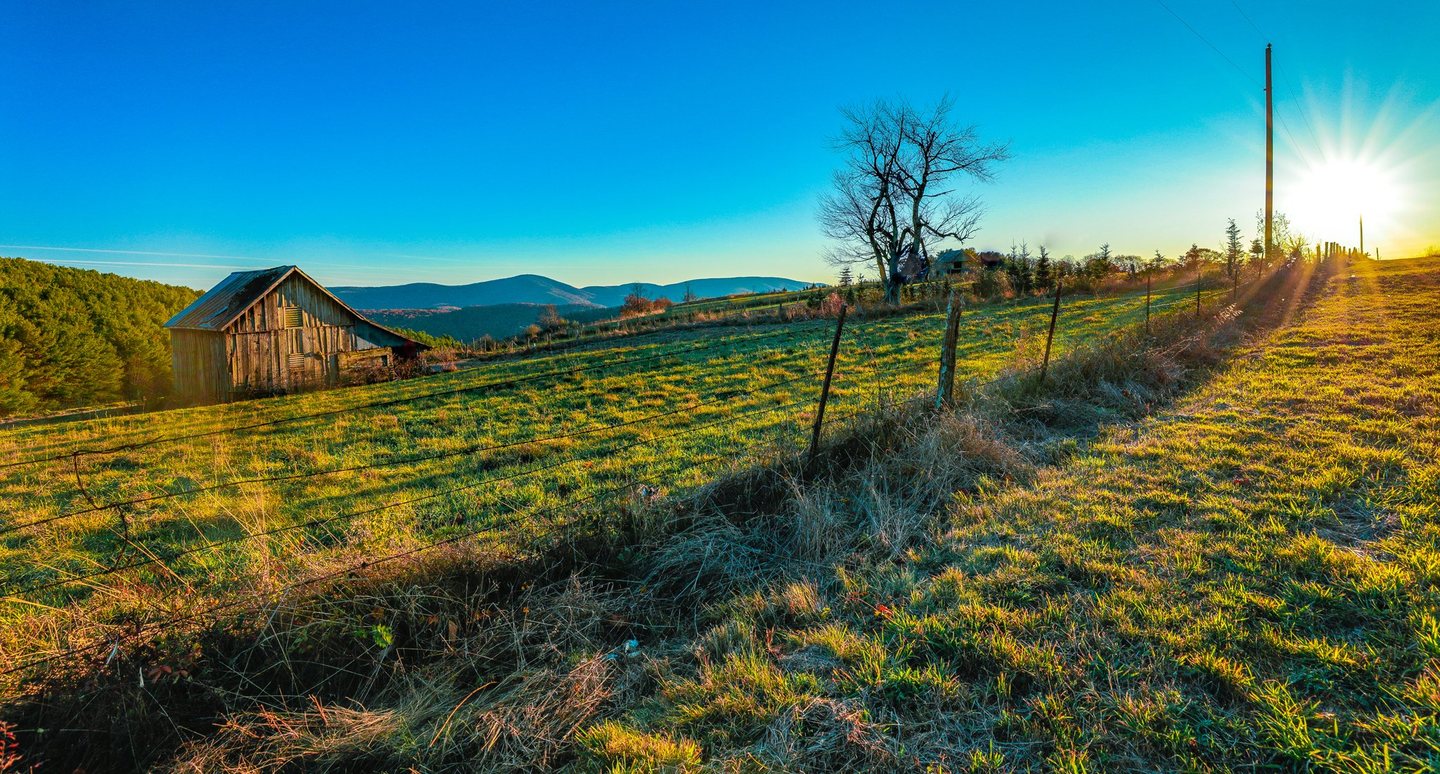 A weathered wooden barn stands in a fenced grassy pasture beneath a clear blue sky, with distant mountain ridges and low sunlight streaming past a bare tree.