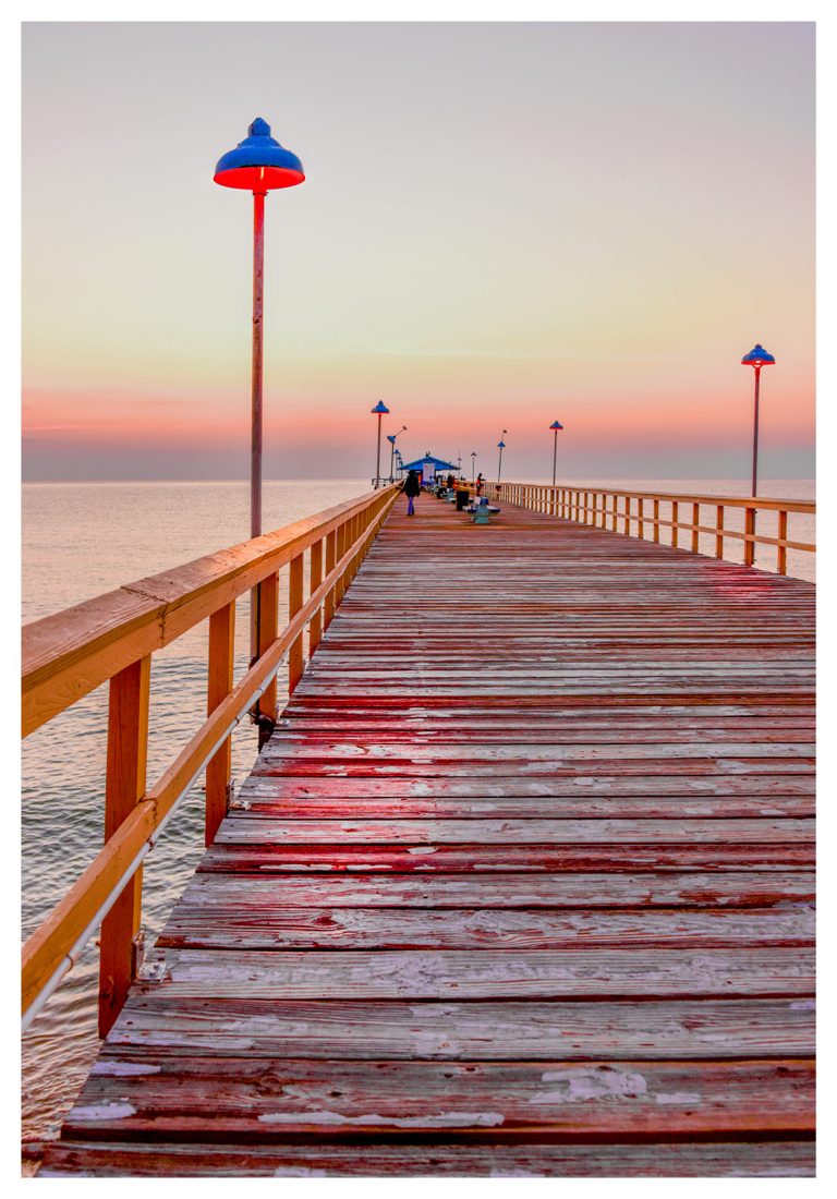A weathered wooden pier lined with blue lamps glowing red stretches across calm water toward a small building and distant people beneath a pastel sky.