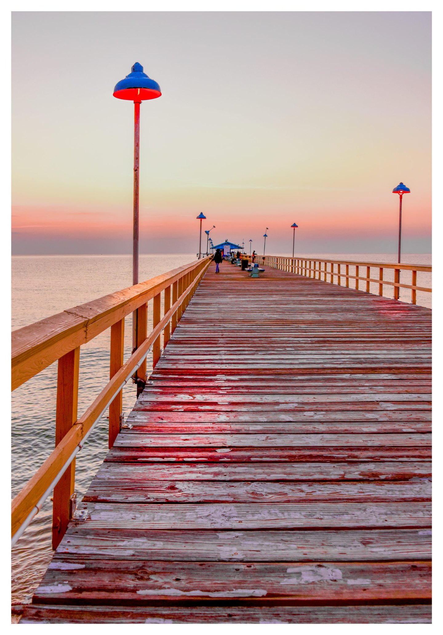 A weathered wooden pier lined with blue lamps glowing red stretches across calm water toward a small building and distant people beneath a pastel sky.