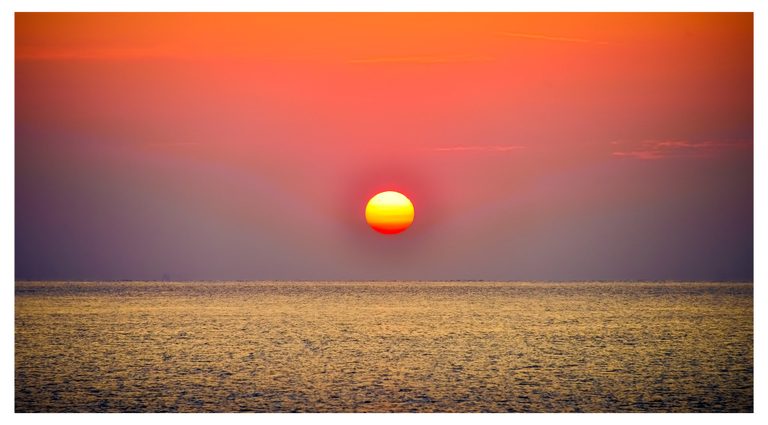 A bright yellow and orange sun hangs above a level sea horizon beneath a coral sky, with golden light across the rippled water.
