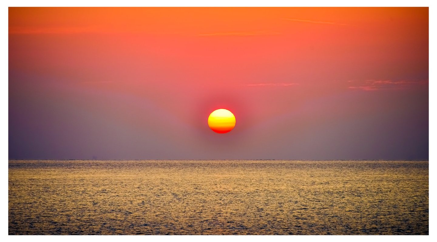 A bright yellow and orange sun hangs above a level sea horizon beneath a coral sky, with golden light across the rippled water.