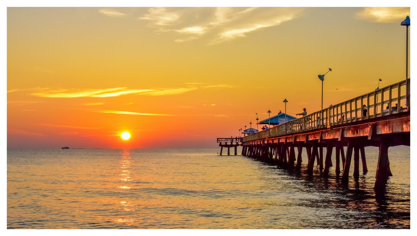 A low sun reflects across calm seawater beneath an orange sky, beside a long fishing pier lined with people and lamps.