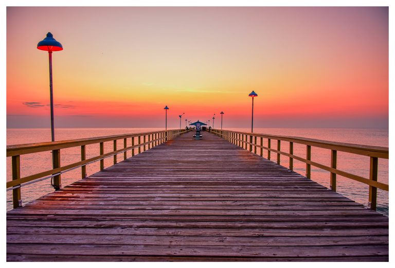 A wooden pier lined with railings and lamps stretches toward a small pavilion across calm water beneath a pink and orange sky.