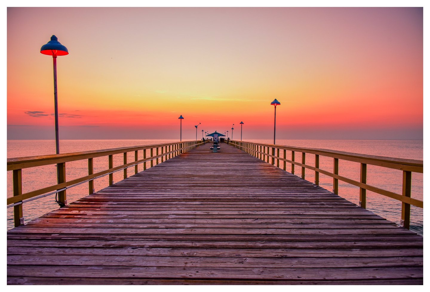 A wooden pier lined with railings and lamps stretches toward a small pavilion across calm water beneath a pink and orange sky.