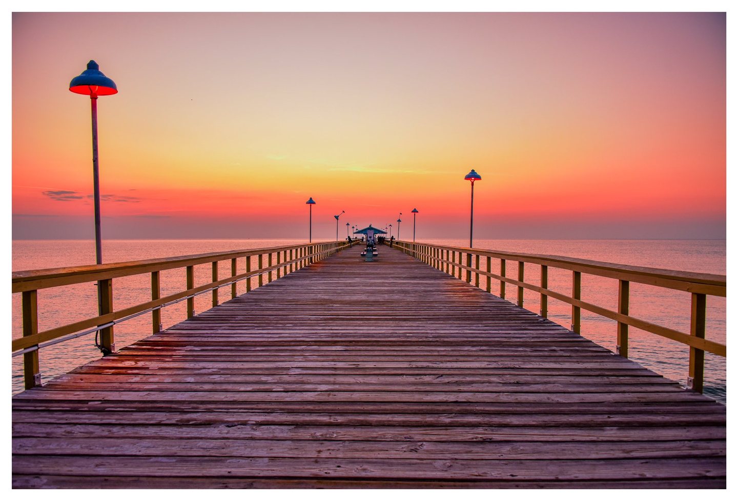 A wooden pier lined with railings and lamps stretches toward a small pavilion across calm water beneath a pink and orange sky.