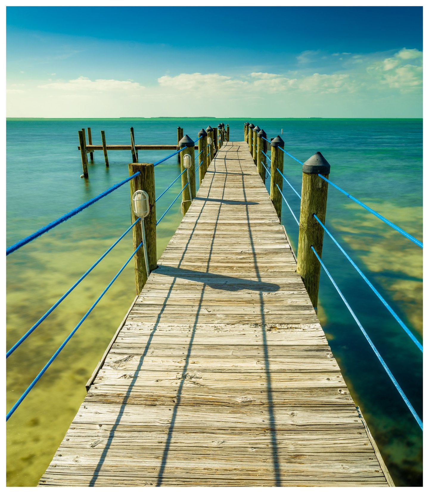 A weathered wooden pier with bright blue rope railings extends over clear turquoise water toward low distant islands beneath a lightly clouded sky.