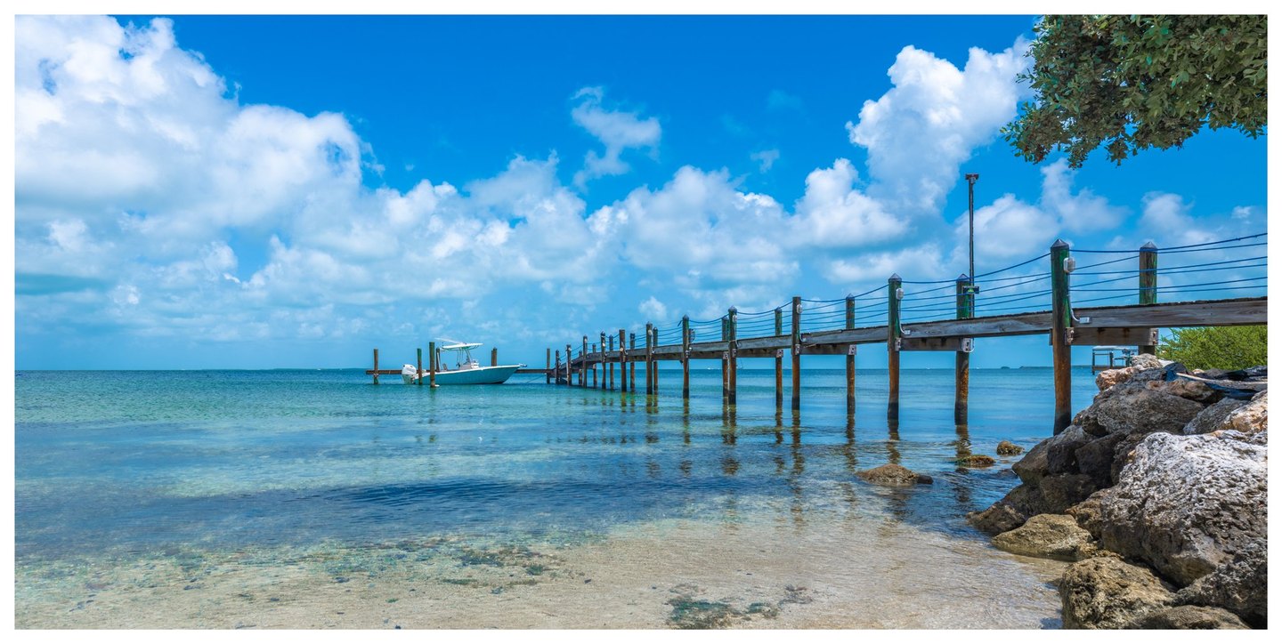 A wooden pier extends from a rocky shoreline across clear turquoise water toward a small motorboat beneath a bright blue sky filled with white clouds.