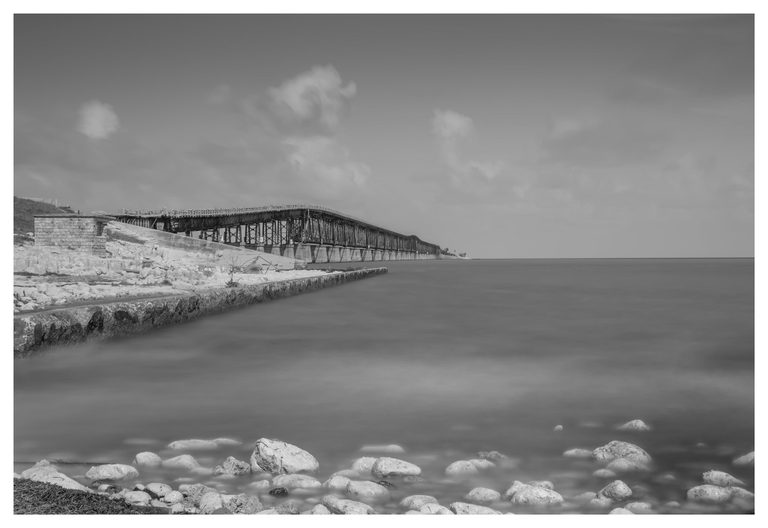 Black-and-white photograph of a long metal bridge receding from a rocky shoreline beside smooth water beneath scattered clouds.