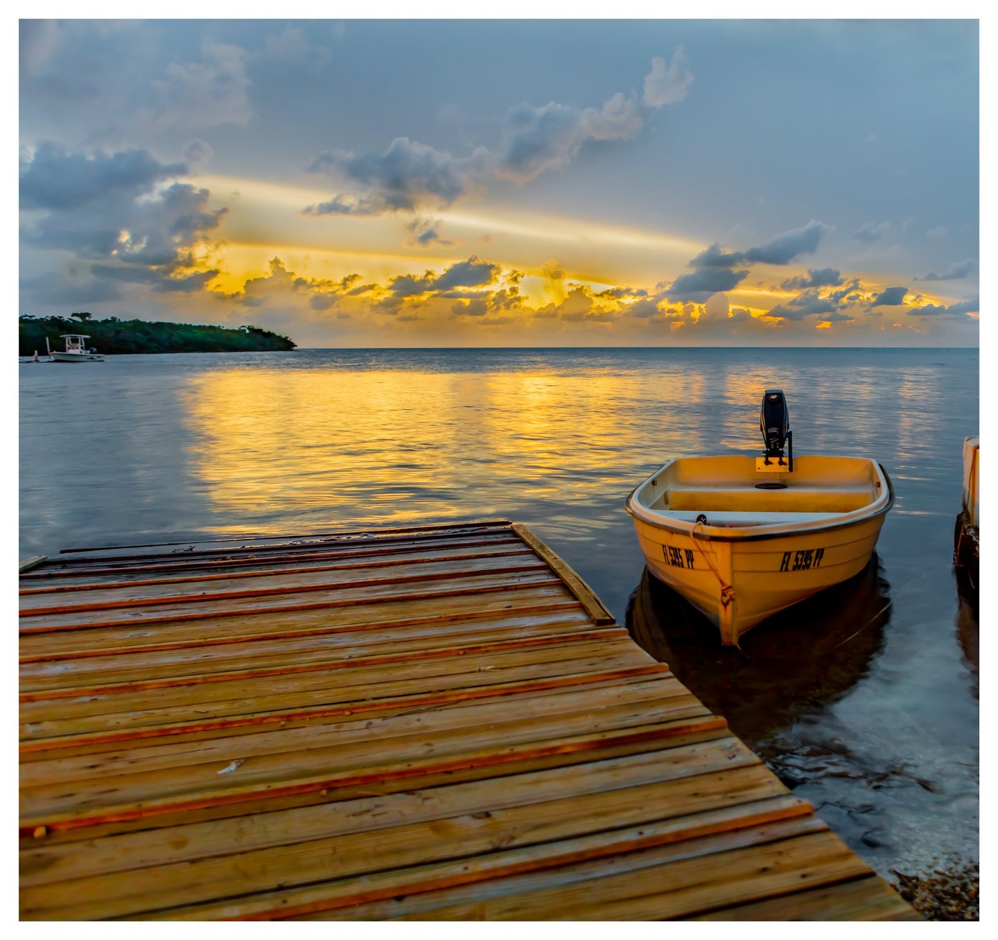 A small motorboat floats beside a wooden dock as golden light breaks through blue-gray clouds and reflects across calm coastal water.
