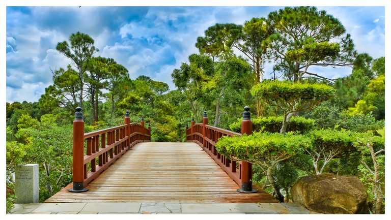 A wooden garden bridge with red railings and black finials leads into dense green trees beneath a blue sky filled with clouds.
