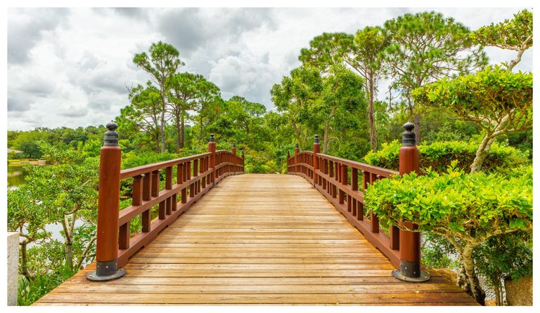 A wooden footbridge with reddish-brown railings crosses a pond amid lush shrubs and tall pine trees beneath a cloudy sky.