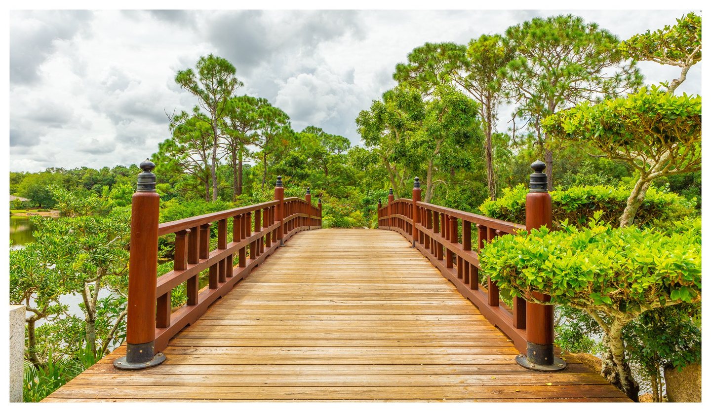 A wooden footbridge with reddish-brown railings crosses a pond amid lush shrubs and tall pine trees beneath a cloudy sky.