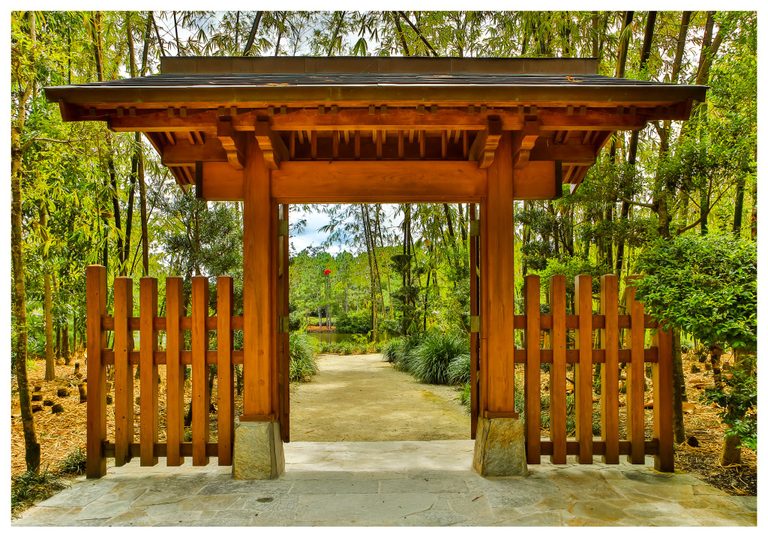 A roofed wooden garden gateway and slatted fence frame a pale path leading through dense bamboo and greenery toward a pond.