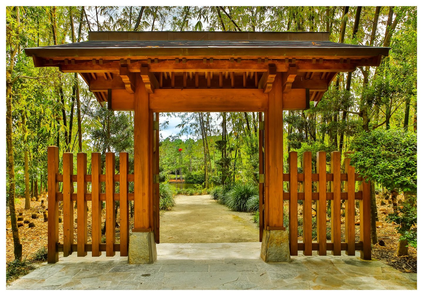 A roofed wooden garden gateway and slatted fence frame a pale path leading through dense bamboo and greenery toward a pond.