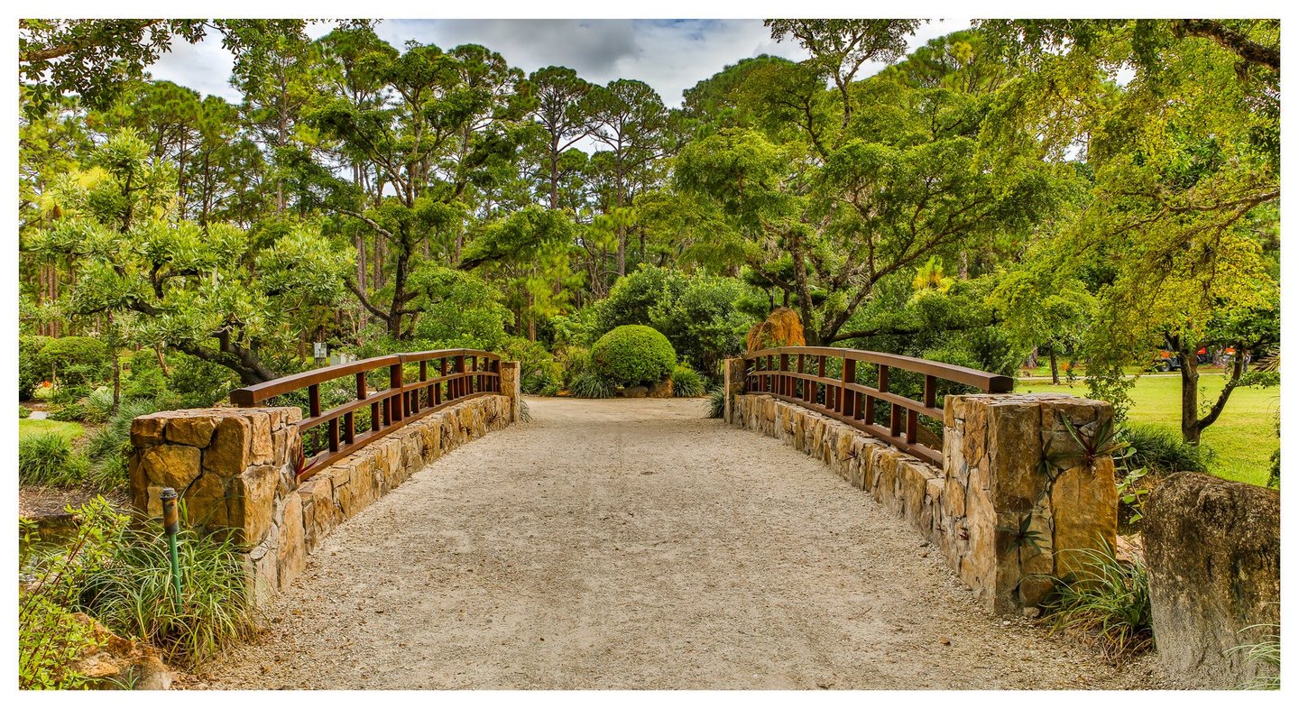 A wide gravel footbridge with stone walls and brown railings leads into a lush garden of rounded shrubs and tall trees.