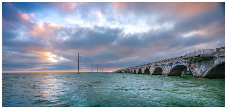 A weathered arched bridge and a parallel line of utility poles stretch across turquoise water toward an orange horizon beneath blue-gray and pink clouds.