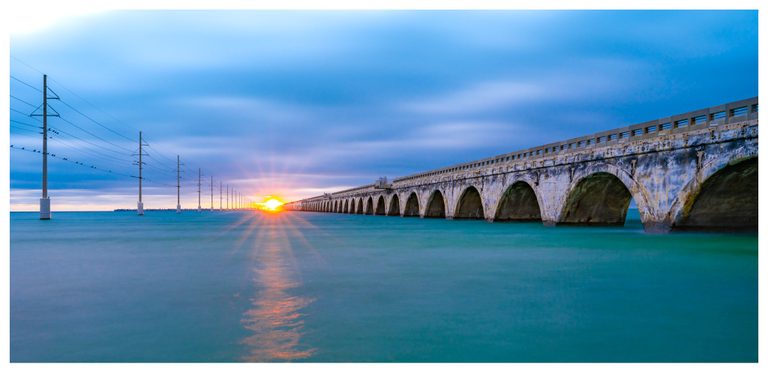 A weathered arched bridge and a parallel line of utility poles stretch across turquoise water toward a low golden sun beneath blue clouds.