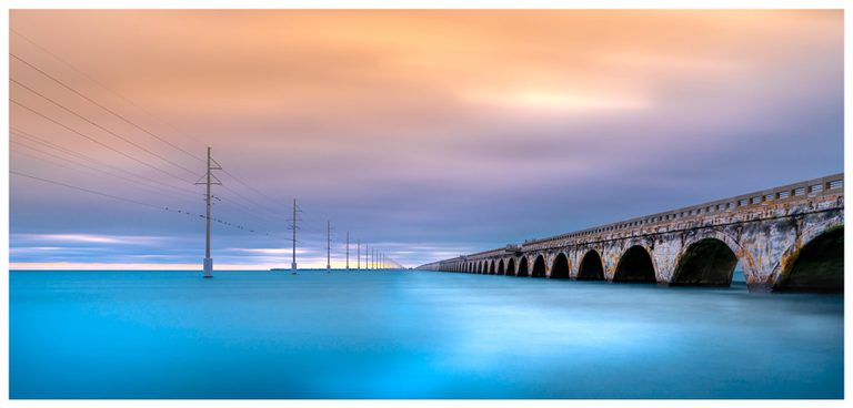 A weathered arched bridge and a parallel line of utility poles recede across turquoise water beneath peach and blue-gray clouds.