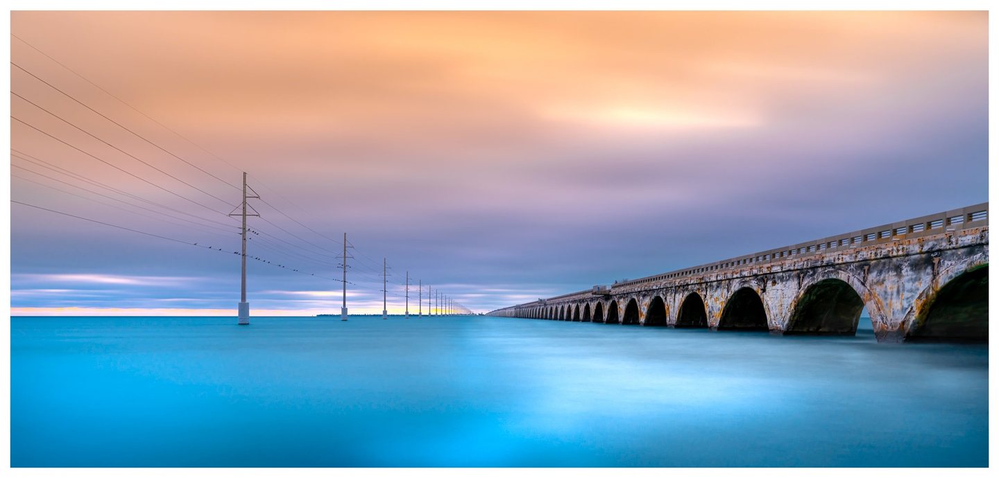 A weathered arched bridge and a parallel line of utility poles recede across turquoise water beneath peach and blue-gray clouds.