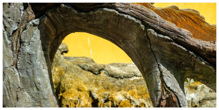A weathered wooden arch with deeply cracked grain frames rough rocks against a bright yellow wall.