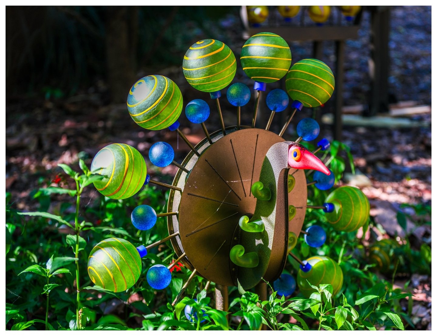 A bird-shaped metal garden ornament with a red beak and a fan of green, yellow-striped globes and smaller blue spheres stands among sunlit plants.