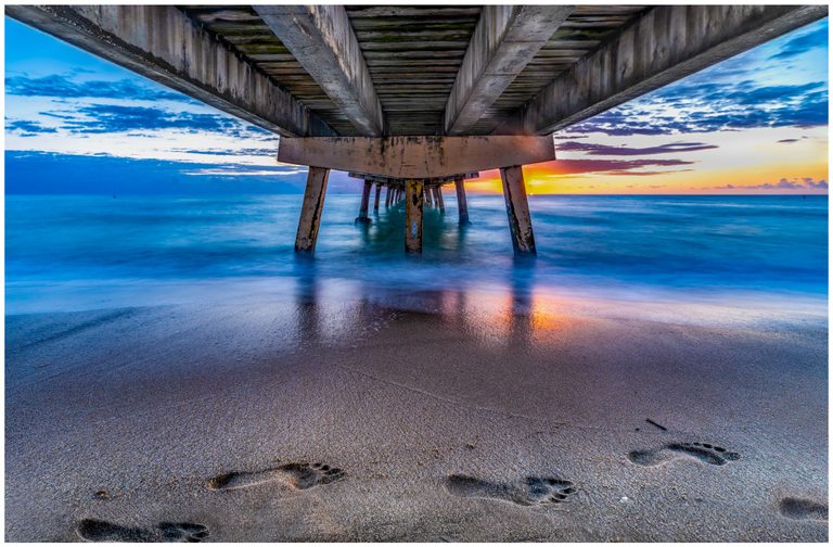 A view beneath a concrete pier shows blue ocean waves, orange light along the horizon, and bare footprints in the foreground sand.