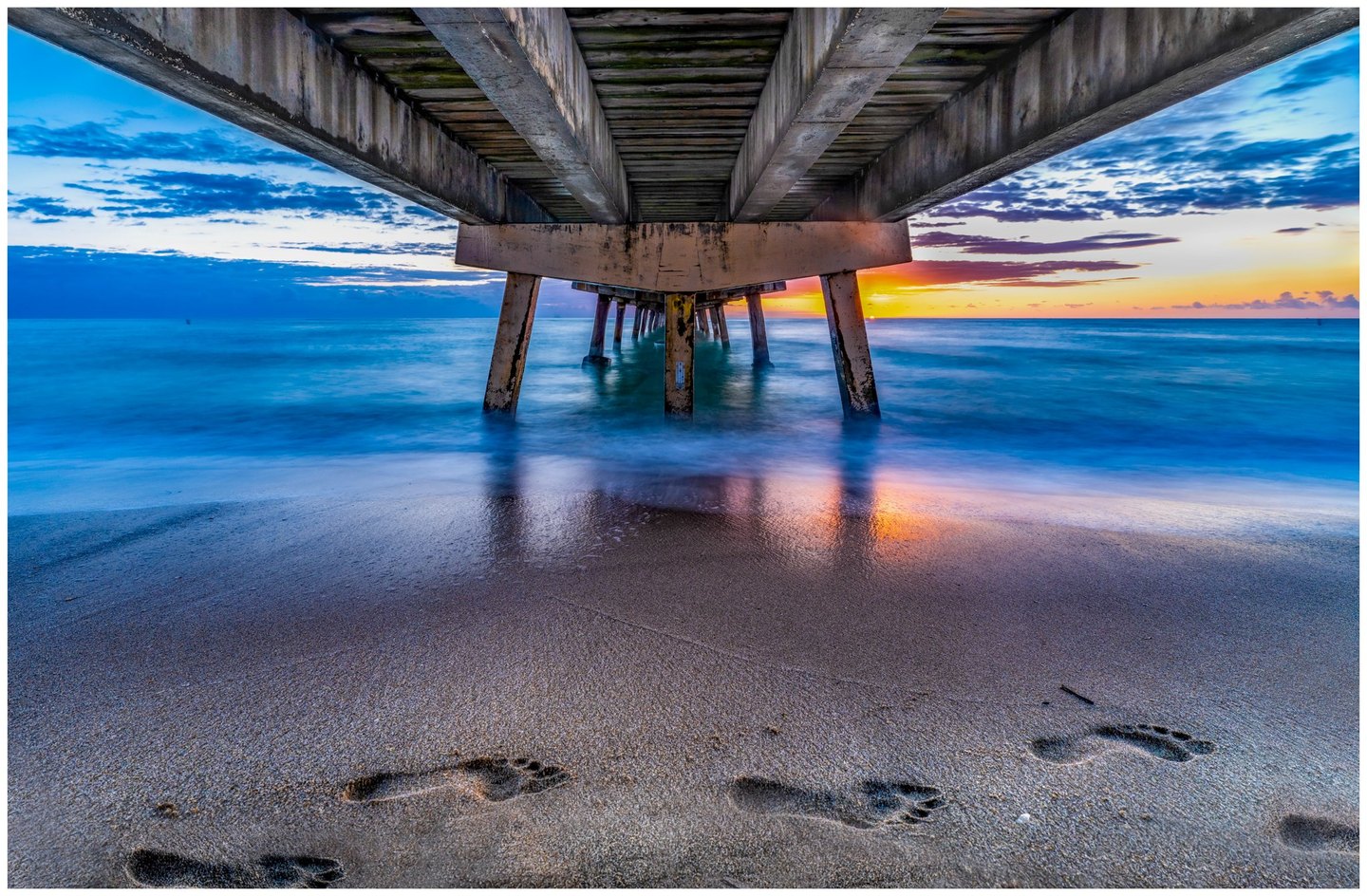 A view beneath a concrete pier shows blue ocean waves, orange light along the horizon, and bare footprints in the foreground sand.