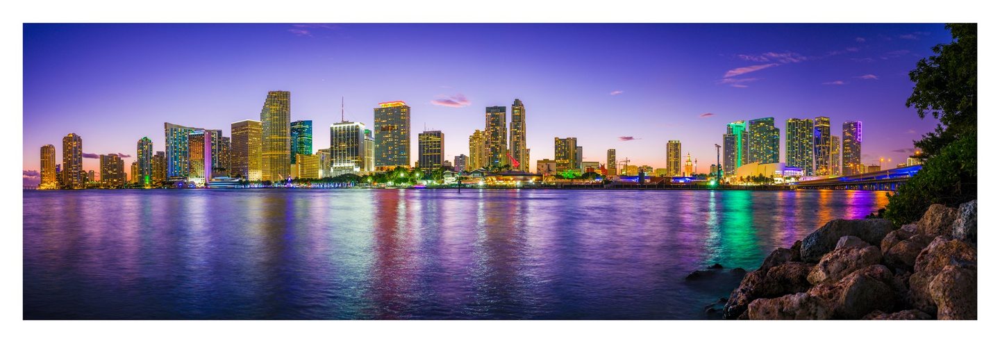 Panoramic view of Miami’s illuminated skyline at twilight, with colorful reflections across the bay and rocks and foliage framing the right foreground.