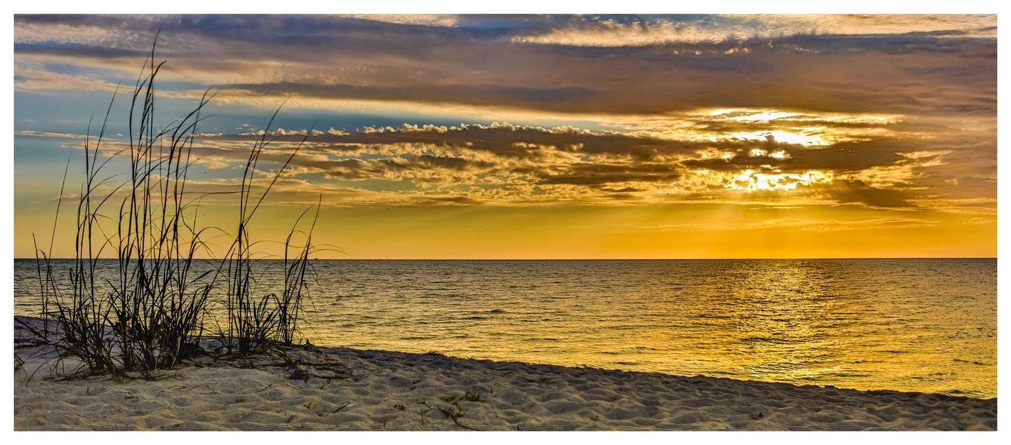 Tall beach grasses rise from a sandy foreground beside calm water, with golden sunlight breaking through layered clouds and reflecting across the surface.