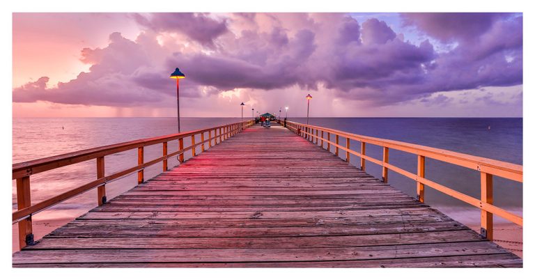 A wooden pier lined with glowing lamps extends into the sea beneath towering pink and purple clouds, with a few people near its far end.