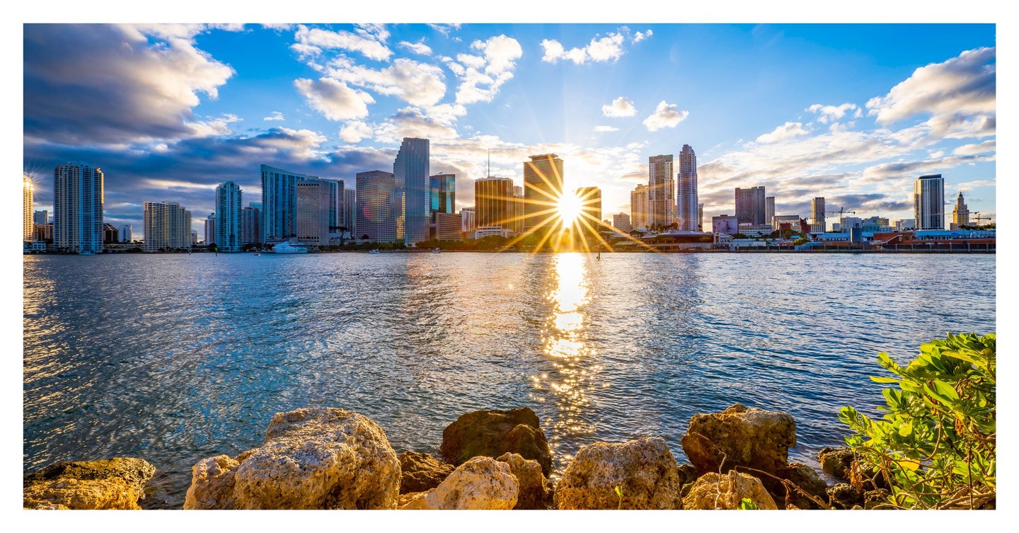 A low sun forms a golden starburst between Miami waterfront towers, reflecting across rippled water toward foreground rocks and green foliage.