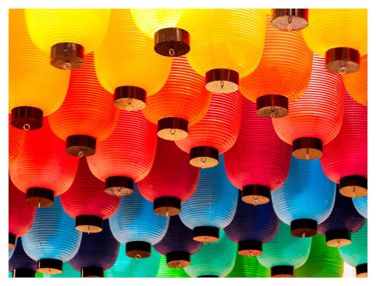 An upward view of densely hung paper lanterns in yellow, orange, red, blue, purple, and green, with ribbed surfaces and dark circular bases.