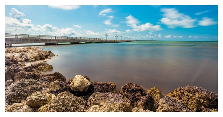 A concrete pier lined with lamps extends from a rocky shoreline across smooth turquoise water beneath a blue sky with streaked white clouds.