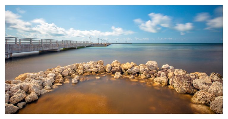 Pale rocks curve around amber shallows in the foreground as a concrete pier lined with lamps extends across smooth turquoise water beneath a blue sky with soft white clouds.