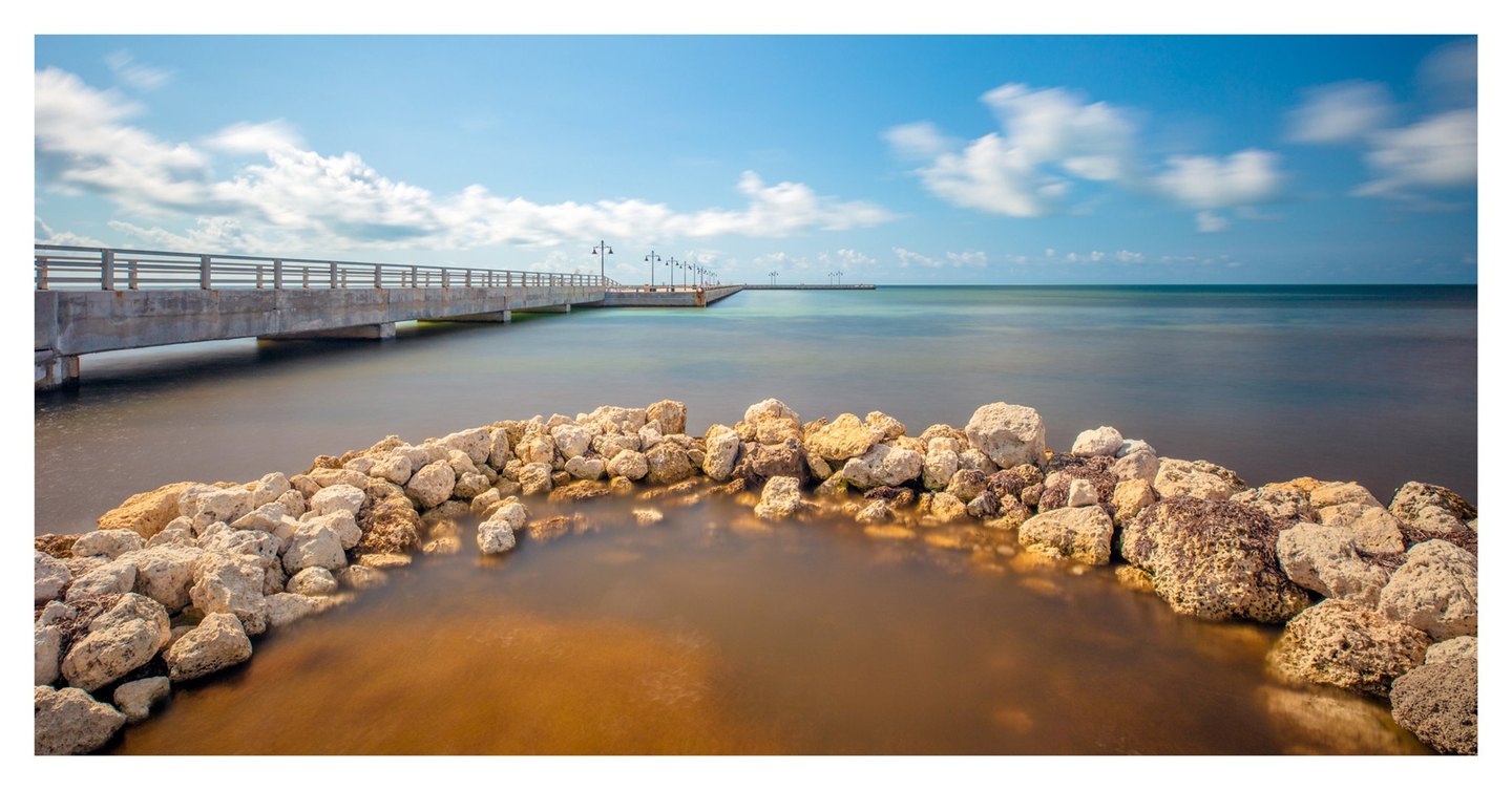 Pale rocks curve around amber shallows in the foreground as a concrete pier lined with lamps extends across smooth turquoise water beneath a blue sky with soft white clouds.