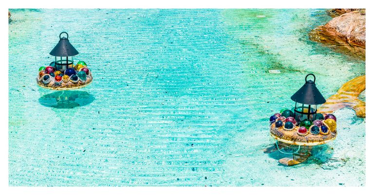 Two black lanterns surrounded by colorful glass spheres on netted circular bases sit in clear turquoise water beside sunlit rocks.