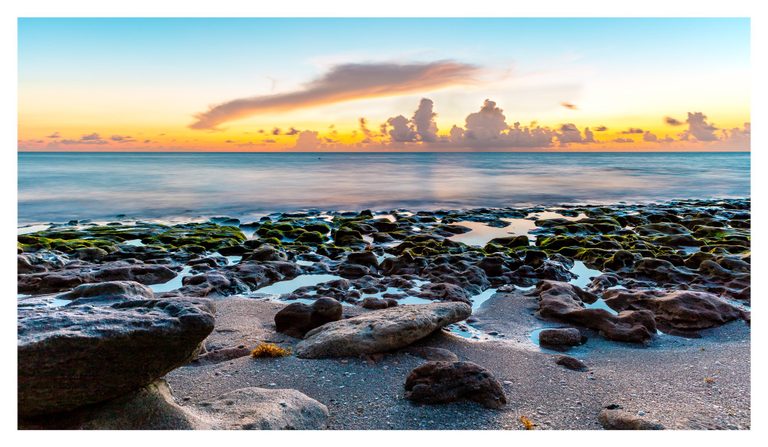 Green algae-covered rocks and reflective tide pools line a sandy shore beneath a turquoise sea and an orange, cloud-filled sky.
