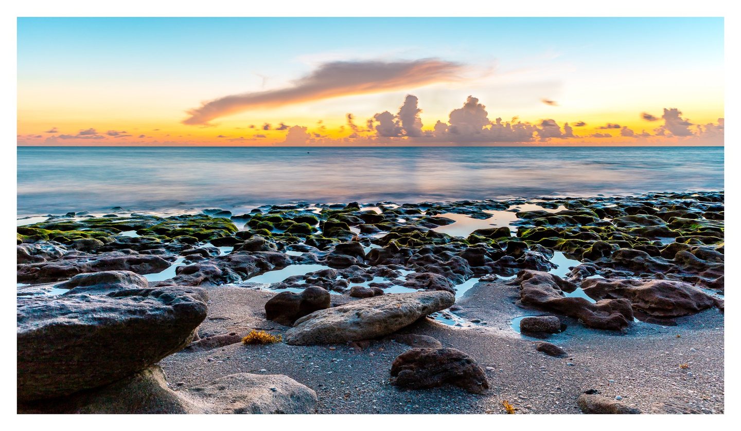 Green algae-covered rocks and reflective tide pools line a sandy shore beneath a turquoise sea and an orange, cloud-filled sky.