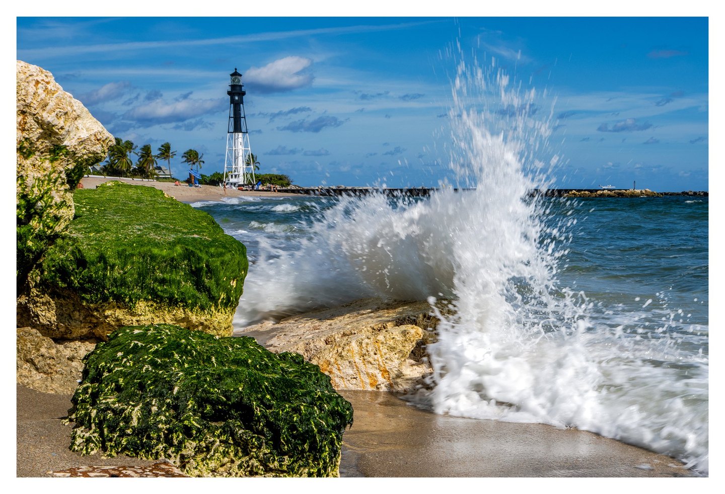 A wave bursts against algae-covered shoreline rocks, with a black-and-white lighthouse and palm trees beyond the sandy beach under a blue sky.